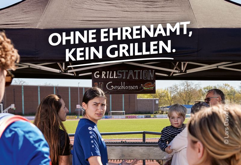 Eine Gruppe Sportlerinnen und Sportler steht vor einer geschlossenen Grillstation auf dem Vereinsgelände. Darüber steht „Ohne Ehrenamt, kein Grillen“. Motiv Grillmeister der LSB NRW Offensive 2026 mit dem Claim „Kein Sport ohne Ehrenamt. Mach Sportehrenamt.nrw“. 