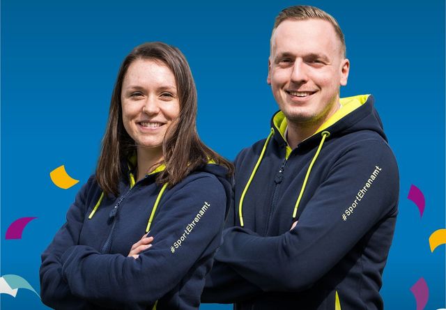 Mann und Frau stehen nebeneinander mit verschränkten Armen. Sie tragen identische dunkelblaue Kapuzenpullover mit gelben Akzenten und gelben Schriftzug „SPORTEHRENAMT“ am Oberarm. Es handelt sich dabei um den #SPORTEHRENAMTShoodie, gesponsert von WestLotto. Konfetti umschwirren die beiden.
