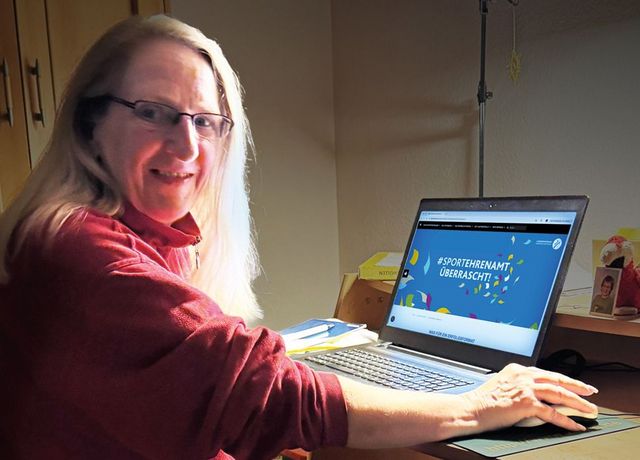 Frau mit rotem Shirt und Brille sitzt früh morgens am Laptop, Bildschirm zeigt #SPORTEHRENAMT ÜBERRASCHT, umgeben von Konfetti