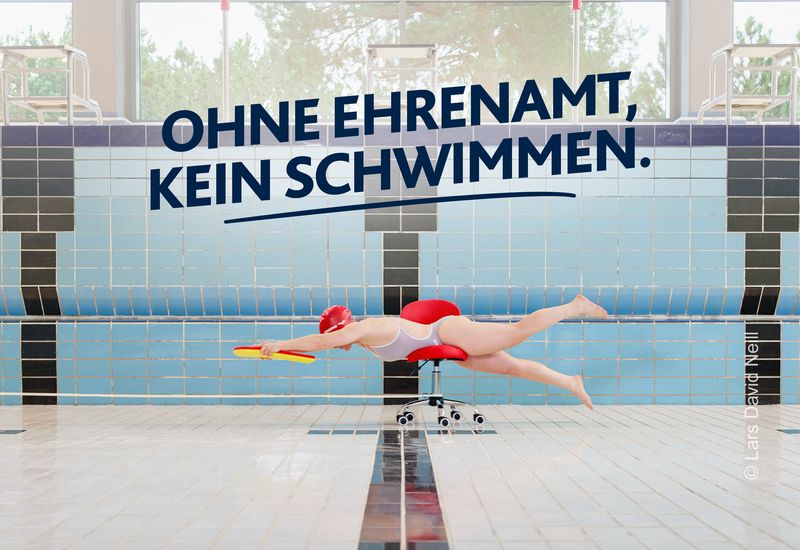 Eine Schwimmerin liegt in Schwimmhaltung auf einem Bürostuhl in einem leeren Schwimmbecken und hält ein gelbes Schwimmbrett. Darüber steht „Ohne Ehrenamt, kein Schwimmen“. Motiv Schwimmen der LSB NRW Offensive 2026 mit dem Claim „Kein Sport ohne Ehrenamt. Mach Sportehrenamt.nrw“.