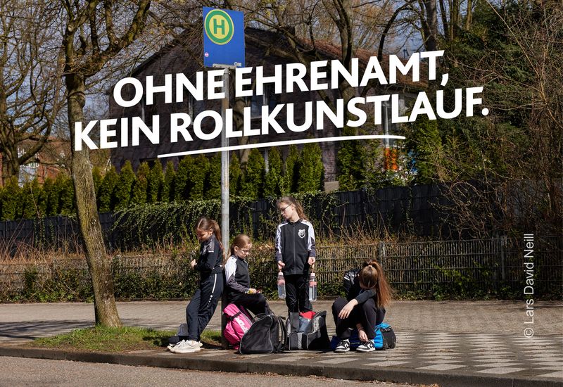 Eine Jugendmannschaft sitzt und steht mit Sporttaschen an einer Bushaltestelle und wartet auf die Fahrgelegenheit zum Training. Darüber steht „Ohne Ehrenamt, kein Rollkunstlauf“. Motiv Elterntaxi der LSB NRW Offensive 2026 mit dem Claim „Kein Sport ohne Ehrenamt. Mach Sportehrenamt.nrw“.
