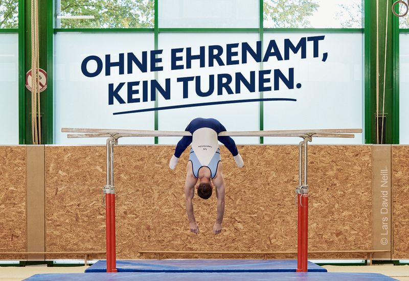 Turner hängt kopfüber mit baumelnden Armen am Barren. Darüber in dunkelblauer Schrift: „Ohne Ehrenamt, kein Turnen.” Motiv der LSB NRW-Offensive 2026: ‚Kein Sport ohne Ehrenamt.‘ 