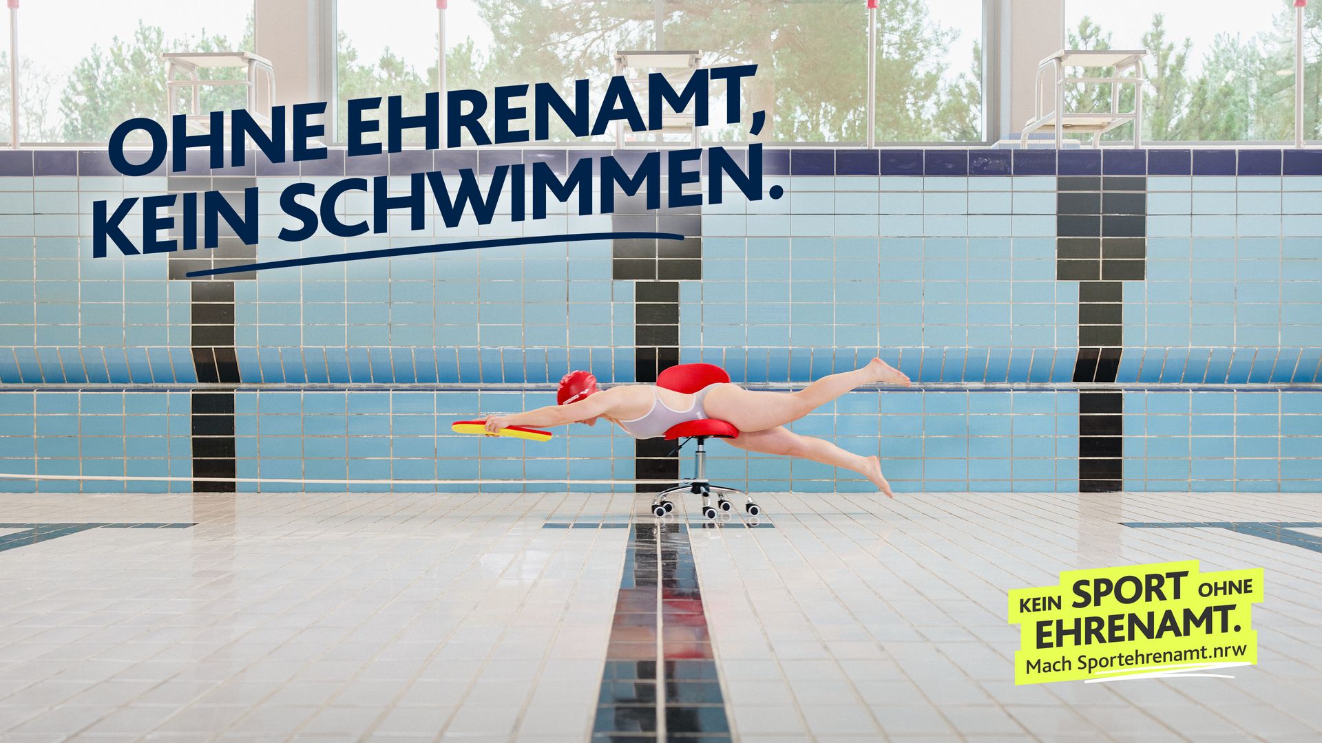 Eine Schwimmerin liegt in Schwimmhaltung auf einem Bürostuhl in einem leeren Schwimmbecken und hält ein gelbes Schwimmbrett. Darüber steht „Ohne Ehrenamt, kein Schwimmen“. Motiv Schwimmen der LSB NRW Offensive 2026 mit dem Claim „Kein Sport ohne Ehrenamt. Mach Sportehrenamt.nrw“.